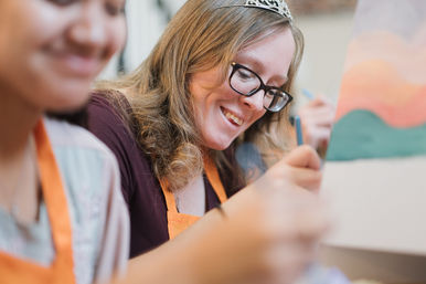 Smiling woman wearing glasses and a tiara painting a colorful canvas at a community art workshop, orange aprons visible