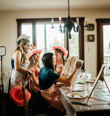 Cheerful indoor painting party in a sunlit dining room, women wearing pink cowboy hats paint on easels at a table with palettes, brushes and drinks.