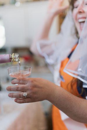 Close-up of rosé being poured into a coupe glass while a laughing person wearing a veil holds it, a sparkling engagement ring visible — joyful celebration scene.