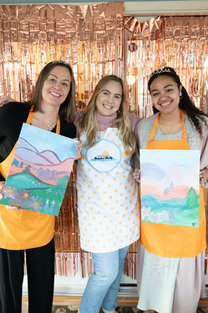 Three smiling women in aprons and tiaras hold colorful landscape canvases at an indoor paint-and-sip party with a rose-gold fringe backdrop.