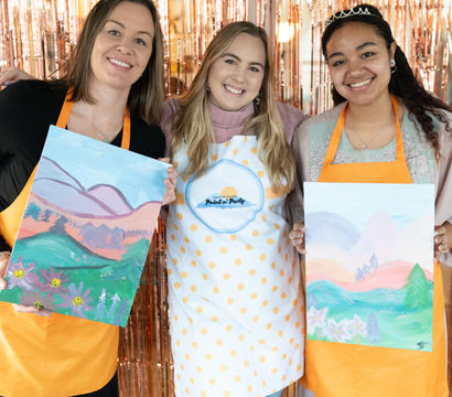Three smiling women in aprons and tiaras hold colorful landscape canvases at an indoor paint-and-sip party with a rose-gold fringe backdrop.