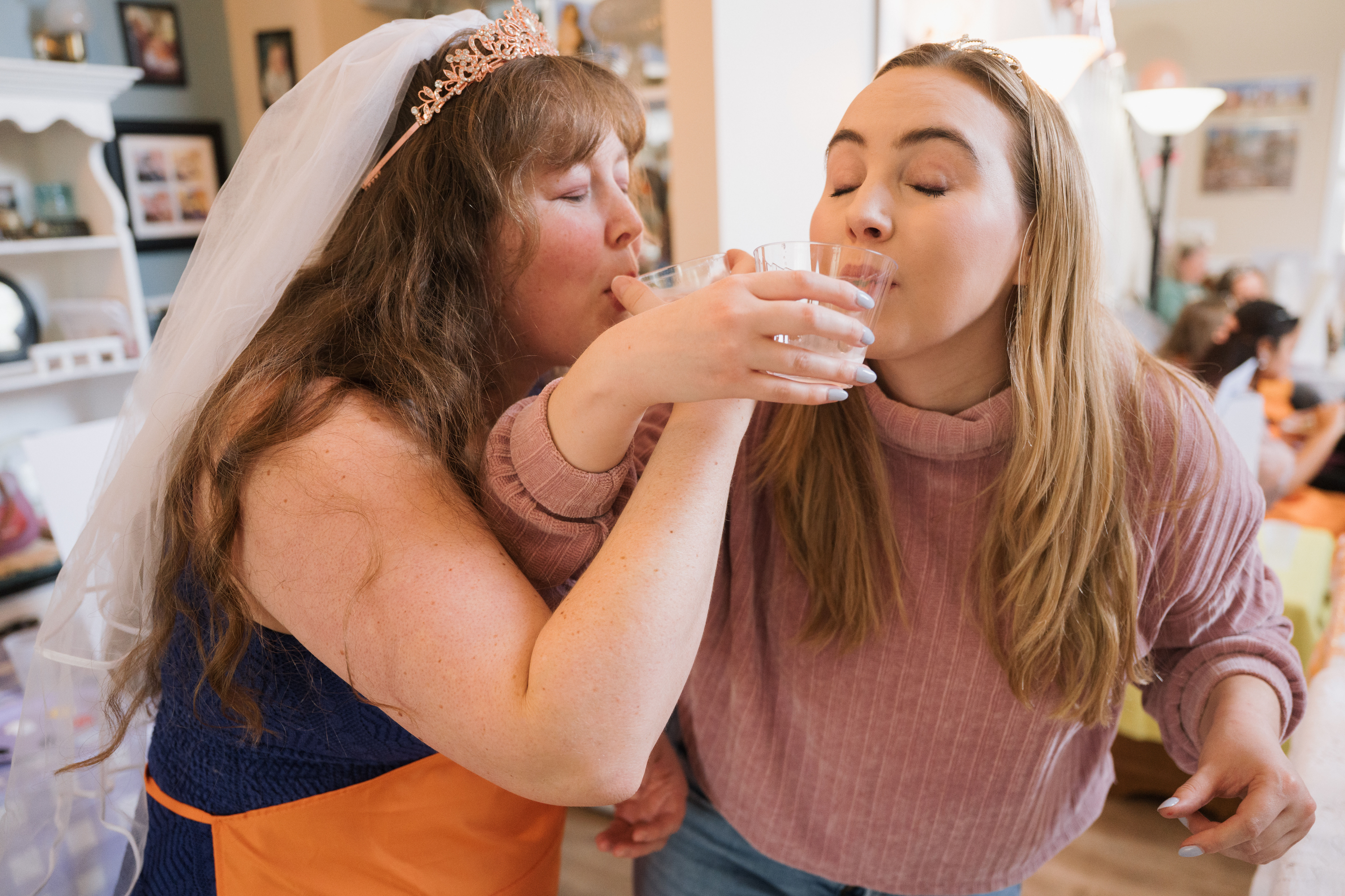 Bride-to-be with veil and friend wearing tiaras sipping from small glasses together at an indoor bachelorette party