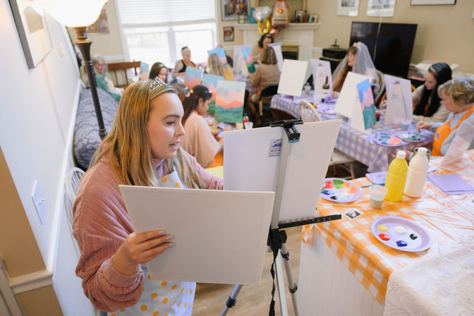Cozy at-home paint night with women in aprons seated at easels and long tables, colorful palettes and canvases on display, foreground woman wearing a small tiara concentrating on her painting.