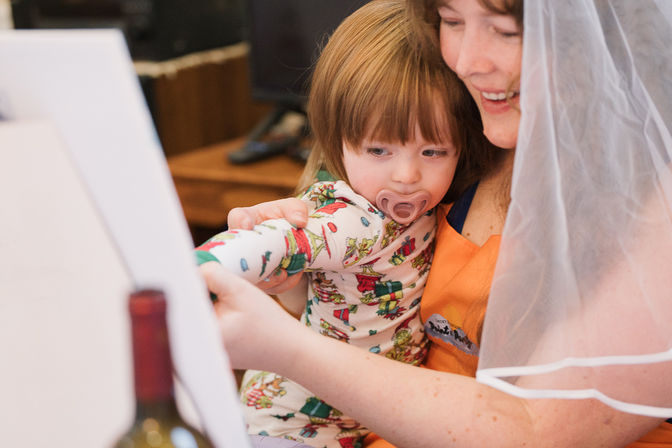 Woman wearing a bridal veil smiles while holding a toddler with a pacifier as they paint together on an easel; child in holiday-print pajamas.