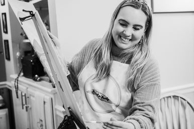 Smiling woman in apron and tiara painting on an easel at an indoor paint-night party