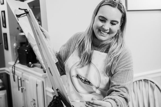 Smiling woman in apron and tiara painting on an easel at an indoor paint-night party