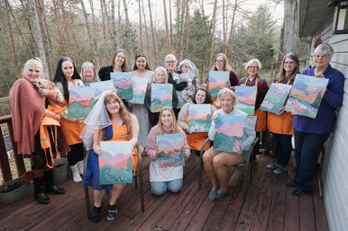 Smiling group of women on a wooden deck in a forested backyard holding pastel mountain landscape canvases at a paint-and-sip party; one wears a bridal veil, a small dog and a child present.