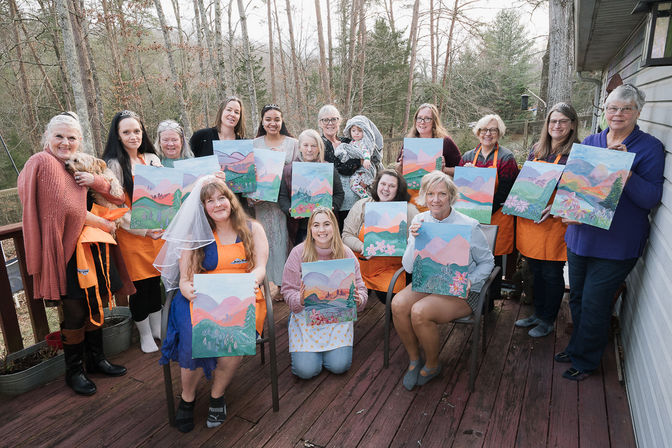 Smiling group of women on a wooden deck in a forested backyard holding pastel mountain landscape canvases at a paint-and-sip party; one wears a bridal veil, a small dog and a child present.