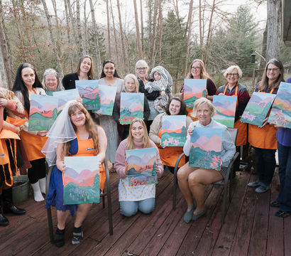 Smiling group of women on a wooden deck in a forested backyard holding pastel mountain landscape canvases at a paint-and-sip party; one wears a bridal veil, a small dog and a child present.