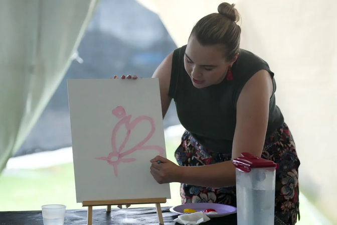 Artist leaning over an easel painting a pink butterfly sketch on a white canvas inside a tented outdoor art workshop, palette and water pitcher on the table.