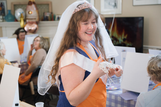 Smiling bride-to-be wearing a veil and tiara at a lively bridal shower painting party, holding a beaded headpiece amid easels and guests.