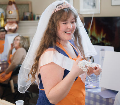 Smiling bride-to-be wearing a veil and tiara at a lively bridal shower painting party, holding a beaded headpiece amid easels and guests.
