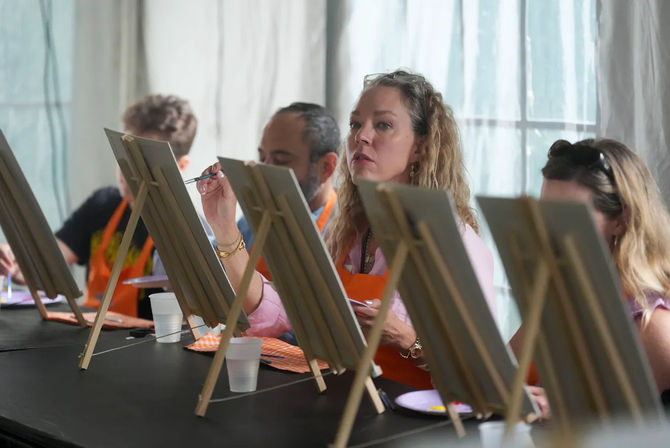 Adults in an indoor group painting workshop seated at easels, a focused woman in an orange apron painting with a brush while paint palettes and plastic cups sit on the table.