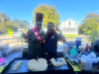 Two people on a sunny residential balcony celebrating a birthday — one wearing a tall red hat and a 'Birthday' sash, the other flashing a peace sign beside a flat-top griddle with rice arranged as 'I ♥ U', with condiment bottles and party supplies nearby.