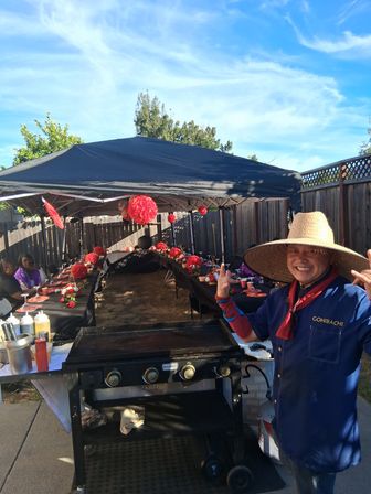 Sunny suburban backyard party with a long table under a black canopy decorated with red paper lanterns and place settings, a flat-top grill in the foreground and a smiling cook in a large straw hat posing beside it.