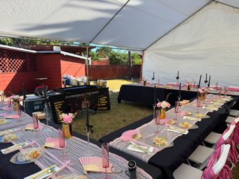 Festive backyard celebration under a white canopy with long black-and-pink tables, floral centerpieces, pink fans, glassware and plated salads on the lawn.