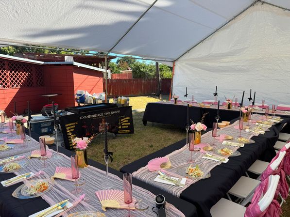 Festive backyard celebration under a white canopy with long black-and-pink tables, floral centerpieces, pink fans, glassware and plated salads on the lawn.