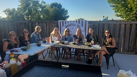 Eight friends wearing sashes gather at a sunny backyard patio brunch, seated around tables with drinks and snacks in front of a striped "Saddlin' Up" banner and wooden fence.