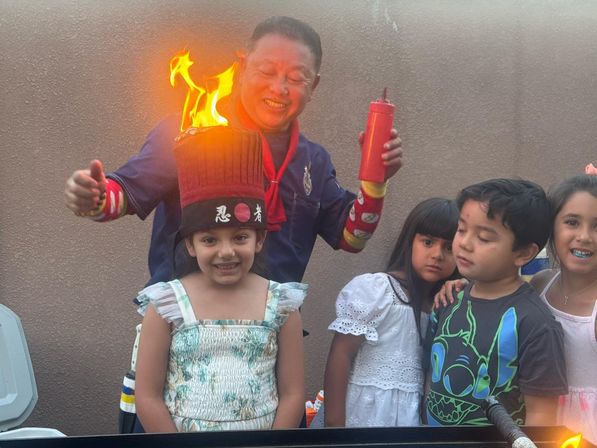 Backyard kids' party: a costumed performer lights a tall novelty hat that bursts into bright flames atop a smiling girl’s head while other children watch nearby.