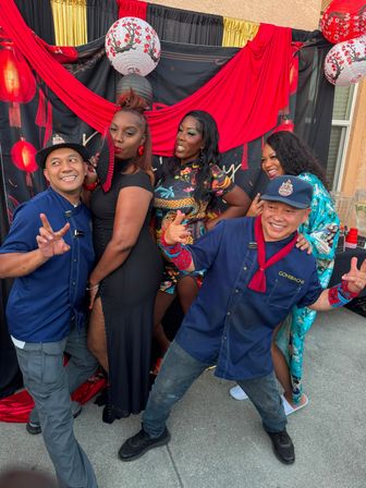 Five people posing playfully at an outdoor Asian-inspired photo booth with red drapery and paper lanterns — two men in blue uniforms, a woman in a black slit dress holding a red fan, and two women in colorful dresses smiling.