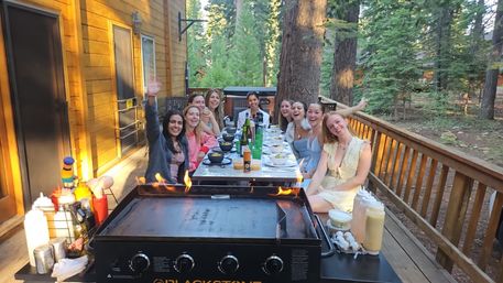 Cheerful group of ten friends gathered at a long table on a wooden cabin deck in a pine forest, flat-top griddle with small flames in the foreground and bowls, bottles and condiments set for an outdoor meal.