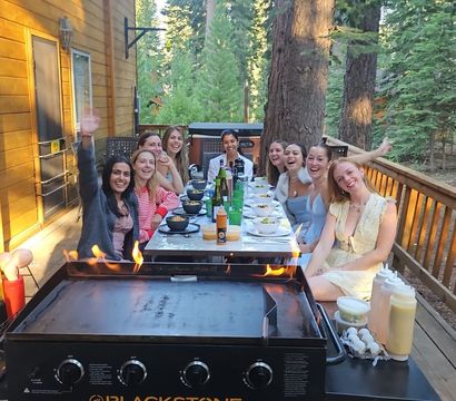 Group of friends laughing and waving around a long outdoor dinner table on a wooden cabin deck in a pine forest, with a flaming griddle in the foreground and bowls, bottles and condiments on the table.