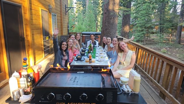 Group of friends laughing and waving around a long outdoor dinner table on a wooden cabin deck in a pine forest, with a flaming griddle in the foreground and bowls, bottles and condiments on the table.