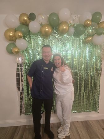 Two people smiling in a party photo in front of a shiny green sequin backdrop with a gold, white and green balloon arch — woman giving a thumbs up at an indoor celebration.