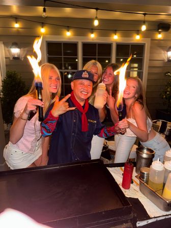 Smiling hibachi chef and four friends holding flaming torches by an outdoor griddle under warm string lights at night