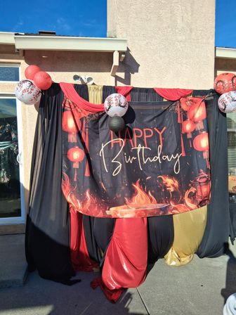 Festive outdoor patio birthday backdrop with red, black and gold drapes, a 'Happy Birthday' banner featuring red paper lanterns and flame graphics, and hanging decorative lanterns.