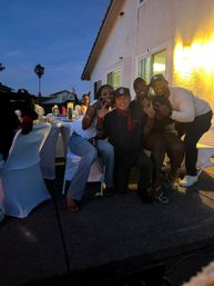 Group of friends posing and flashing peace signs at an evening backyard patio dinner party with white-covered chairs, lit house wall and a palm tree silhouette