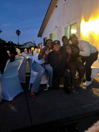 Group of friends posing and flashing peace signs at an evening backyard patio dinner party with white-covered chairs, lit house wall and a palm tree silhouette