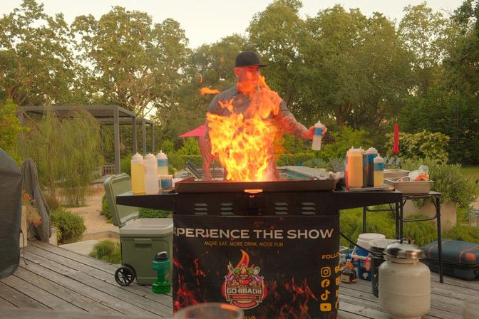Person cooking at an outdoor hibachi-style grill with dramatic orange flames, squeeze bottles of sauces and a propane tank on a wooden backyard patio surrounded by trees.