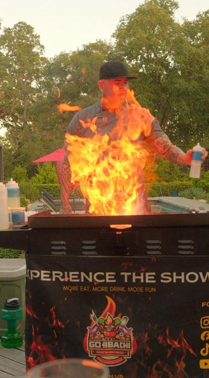 Person cooking at an outdoor hibachi-style grill with dramatic orange flames, squeeze bottles of sauces and a propane tank on a wooden backyard patio surrounded by trees.