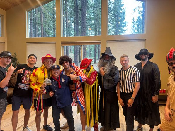 Group of men in colorful costumes posing inside a wood-paneled mountain lodge with tall windows overlooking a pine forest — cowboy hats, star-spangled jacket, wizard robes, referee shirt, and novelty hats at a festive indoor costume party.