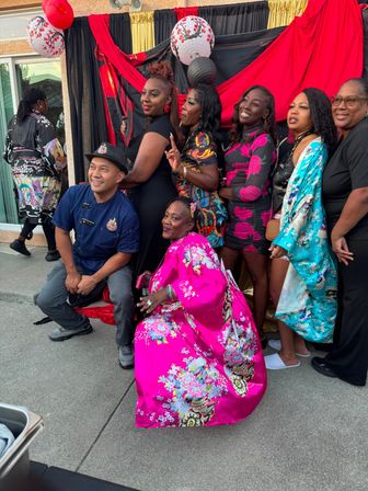 Group of adults posing at a backyard party in front of a red-and-black draped backdrop with paper lanterns, wearing bright floral kimonos and patterned dresses, smiling and striking playful poses.