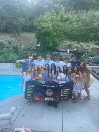 Smiling group of teens and a chef posing with a promotional banner at a backyard pool party, surrounded by succulents, terraced hillside and trees in a sunny outdoor setting