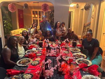 Family birthday dinner party in a home dining room — long table with red and black linens, cherry blossom garlands and lantern centerpieces, plates of food and smiling guests posing together.