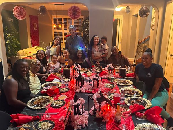 Family birthday dinner party in a home dining room — long table with red and black linens, cherry blossom garlands and lantern centerpieces, plates of food and smiling guests posing together.