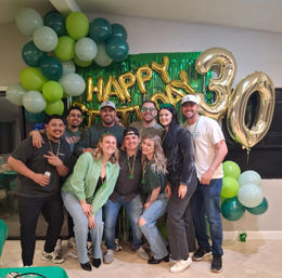 Group portrait of smiling adults at an indoor green-and-gold 30th birthday party with large gold '30' balloons, 'Happy Birthday' letters, green fringe backdrop and balloon garland.