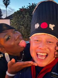 Playful backyard selfie at dusk — smiling person wearing a tall black hat with Japanese characters and a red circle, friend leaning in and sticking out their tongue.
