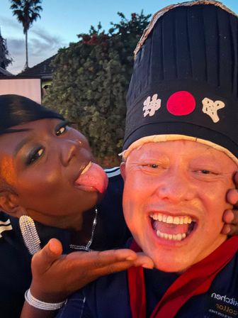 Playful backyard selfie at dusk — smiling person wearing a tall black hat with Japanese characters and a red circle, friend leaning in and sticking out their tongue.