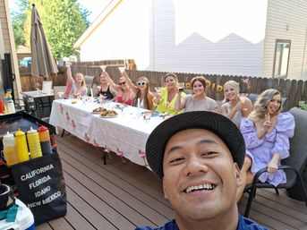 Selfie of a smiling man in front of a lively backyard deck party — a long, flower‑decorated table with eight women raising pink cocktails, a platter of food, and a grill with condiment bottles nearby.