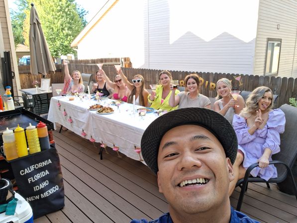 Selfie of a smiling man in front of a lively backyard deck party — a long, flower‑decorated table with eight women raising pink cocktails, a platter of food, and a grill with condiment bottles nearby.
