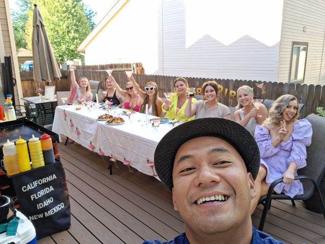 Smiling selfie at a sunny backyard deck party with a group of friends seated at a long decorated table, raising pink cocktails above plates of food on an outdoor patio.