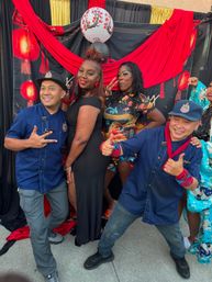 Group photo at an outdoor Asian lantern–themed backdrop with red drapes: two men in blue shirts and caps giving thumbs-up and two women in black and floral dresses posing playfully.