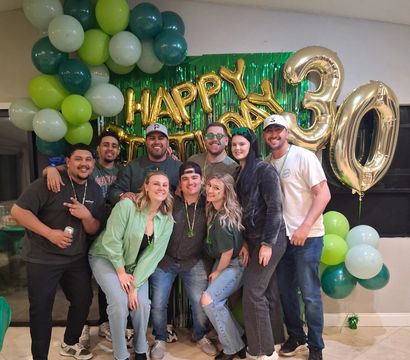 Group of friends posing indoors at a green-and-gold 30th birthday party with balloon garlands, foil "Happy Birthday" backdrop and large gold "30" balloons.