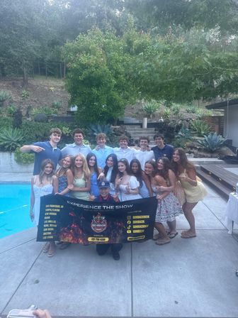 Smiling group at a backyard poolside gathering holding a black promotional banner on a concrete patio, surrounded by succulents, stairs and hillside landscaping.