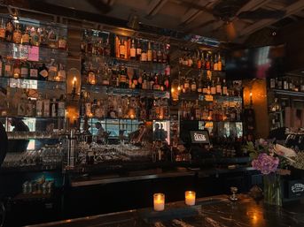 Moody downtown bar interior with backlit liquor shelves, rows of glassware, lit candles on a marble counter and a vase of flowers.