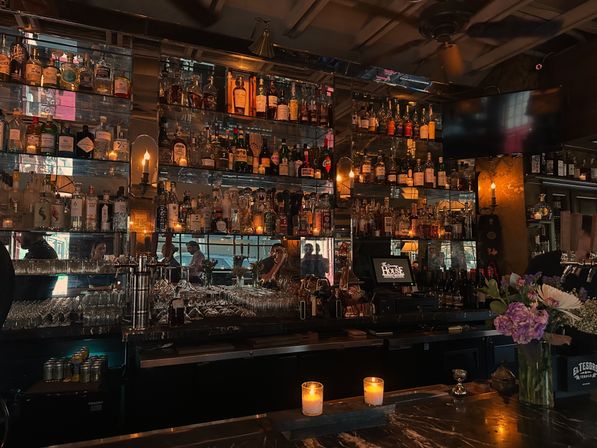 Moody downtown bar interior with backlit liquor shelves, rows of glassware, lit candles on a marble counter and a vase of flowers.